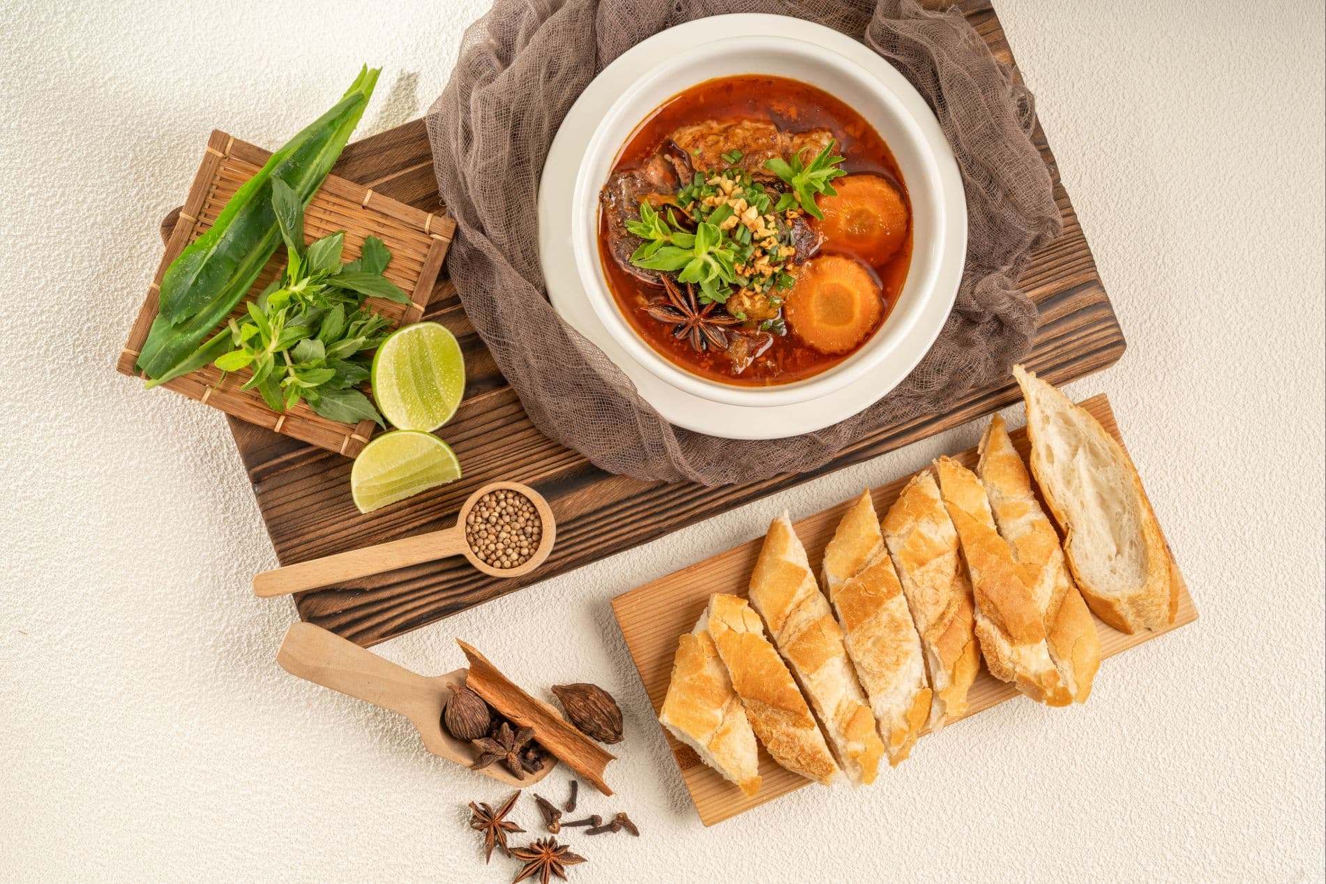 Khmer beef stew photographed from above, showing soft beef, carrot rounds and star anise in red broth, with a board of sliced baguette, lime halves and cinnamon alongside