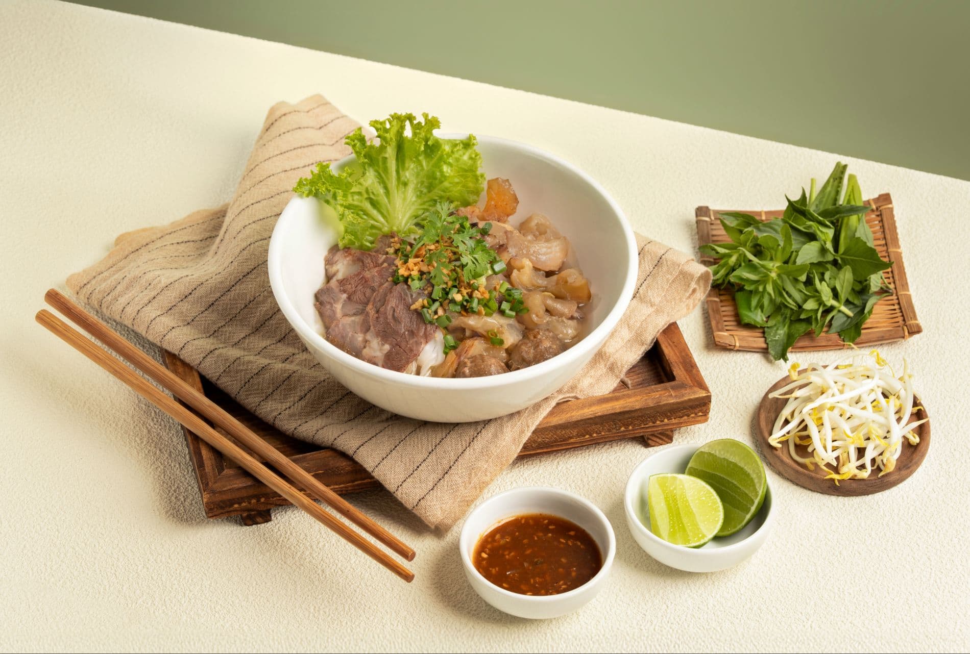 A deep white bowl of beef tendon noodle soup on a wooden stand, with chopsticks, a dish of chilli sauce, lime wedges, bean sprouts and a plate of fresh herbs arranged around it