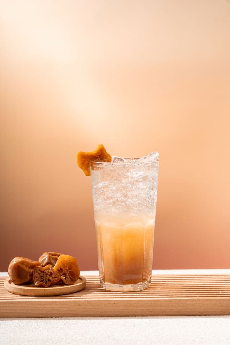 A tall glass of Ngam Ngov preserved-lime soda over ice, garnished with a dried preserved lime, with more preserved limes on a small wooden plate