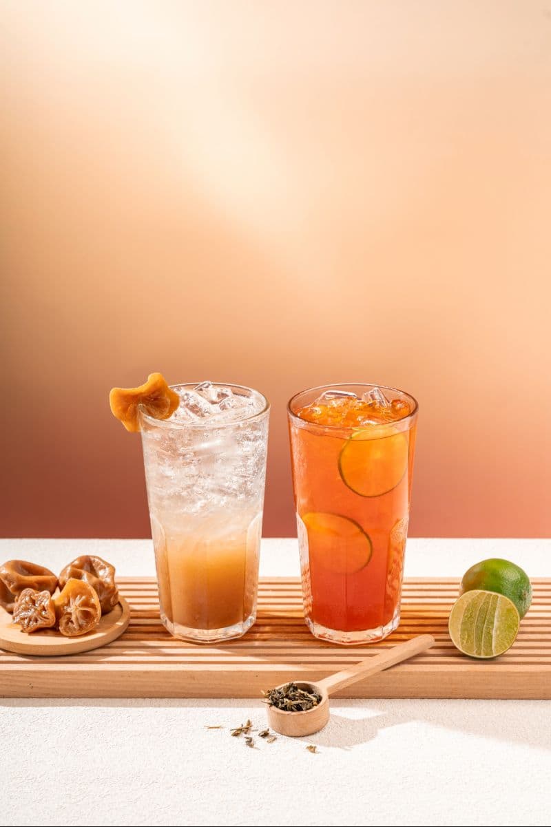 Two cold drinks side by side on a wooden board: a Ngam Ngov preserved-lime soda and an iced lemon tea with lime slices, next to loose tea leaves and a halved lime